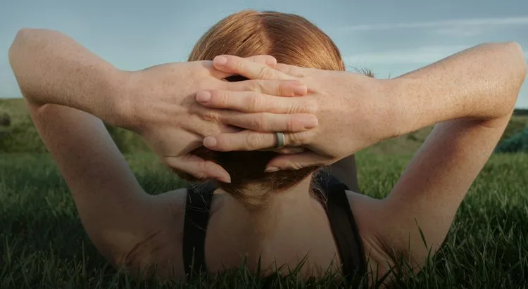 A woman wearing an Oura Ring lies on the grass with her hands clasped behind her head.